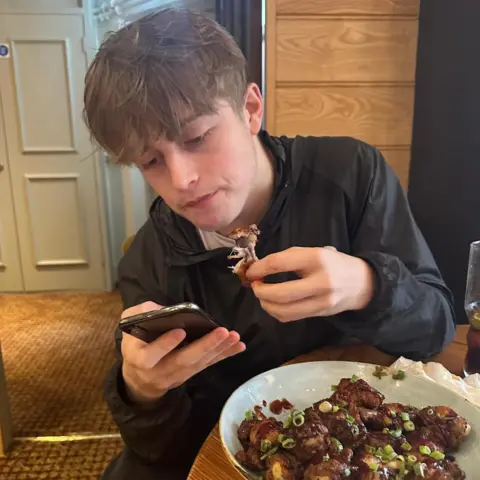Martell Blackford Jacob Holman in a candid picture. He is a young boy with a short back and sides style haircut. He is looking at his phone and eating a plate of chicken wings. 