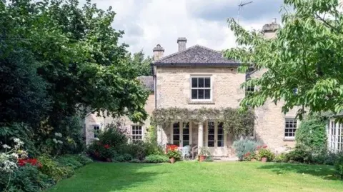 Getty Images A large stone house with two doors leading to a garden with grass and trees along the side. There are red and white flowers in the beds beside the grass.