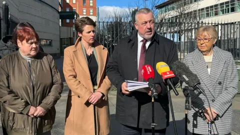 Four people standing outside the Belfast Crown Court. A person wearing a coat, shirt and tie spoke on the microphone holding the notepad. There are three women standing on both sides