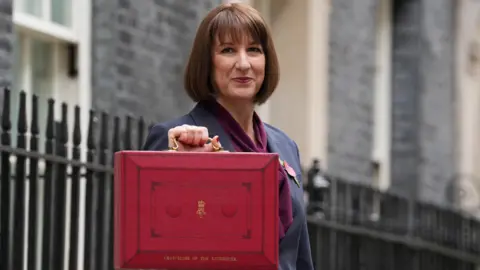 Rachel Reeves, a woman with dark brown hair, dressed in a grey suit with purple scarf, holds up a red box with gold coloured handle and gold coloured lettering in street with black railings behind her.