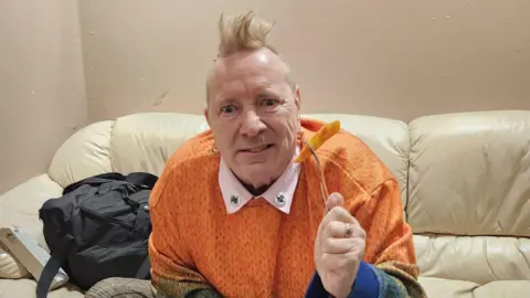 John Lydon An older man, John Lydon, sitting on a cream leather sofa, he is wearing an orange jumper and holding up a fork with an orange chip on. He has a blonde mohawk and is looking into the camera.