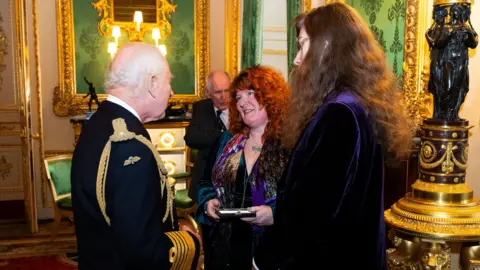 PA Media King Charles III presents an Elizabeth Emblem to Rebecca Lombard-Earl. They are in a gold and green grandly decorated room. Her son Rowan is also with her,