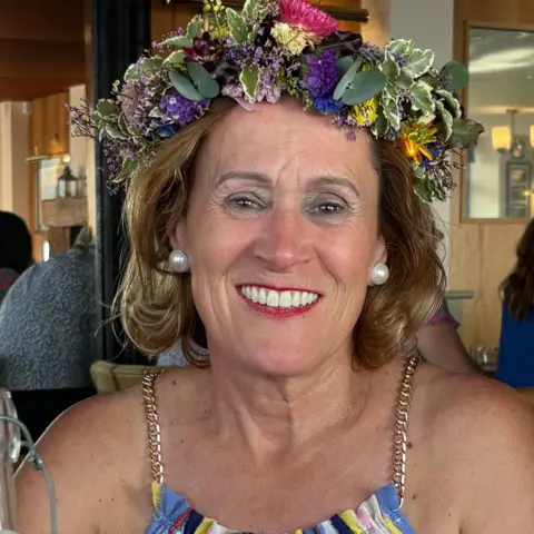 Alice de Freitas Alice de Freitas is wearing a floral garland on her head with blue, yellow and pink flowers. She has a multicoloured striped top on too.