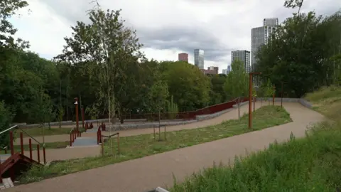 A winding path stone path leads through an area of new plants, trees and shrubs. In the background a number of tower blocks can bee seen behind more established trees. There is also stone steps with red metal railings.