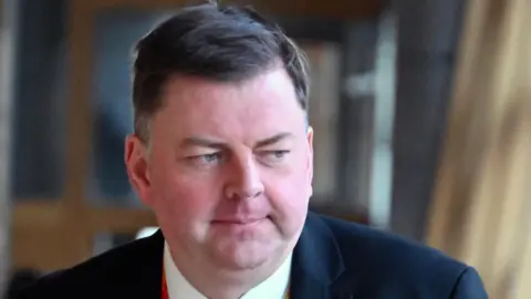 Getty Images A man with short dark hair, wearing a dark suit, white shirt and a lanyard. He is visible from the shoulders up, with dimly lit walls behind him.