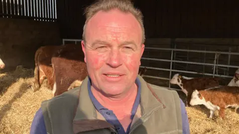 Martin Giles/BBC Chris Blaxell wears a blue polo top and a green gillet. He is standing in a cattle barn with cows behind him and the floor is covered in hay. 