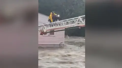 A man and woman crawl on a fire engine ladder above rushing water