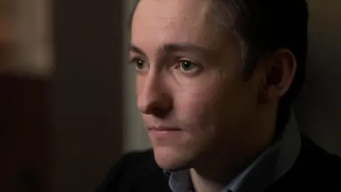 Channel 4 A man with dark brown hair wearing a blue shift and dark jumper. 