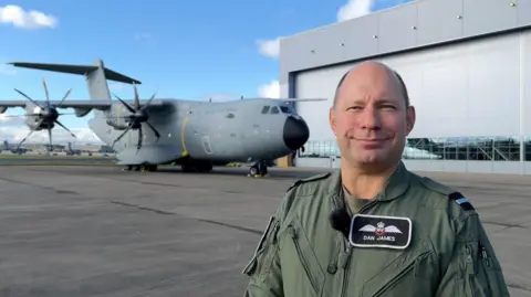 Air Commodore Dan James in his flight suit stands in front of the plane outside a hangar.