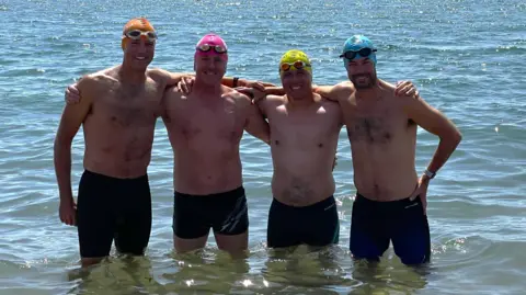 Four men stand in the shallows of the sea. They are all smiling at the camera. They are wearing different colour swim hats - orange, pink, yellow, and blue from left to right.