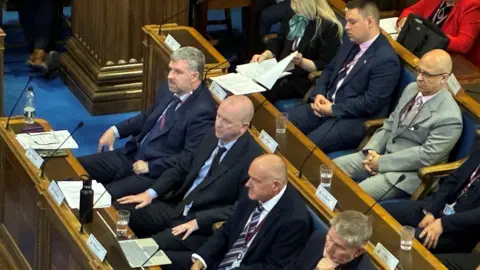 BBC/Lucy Ashton A row of men in navy suits are sitting at wooden desks in a council chamber