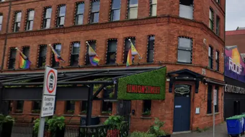 Google Maps A photo of a bar from the street. A sign decorated with artificial grass states UNION STREET. The building is red brick and several rainbow flags are on masts beside windows. Some bins are on the street and some cars are parked up the street. 