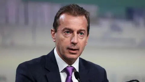 Guillaume Faury is pictured during a press conference. He is speaking into a small black microphone. He is dressed in a navy blue suit, a white shirt and a purple tie. The background is completely out of focus. 