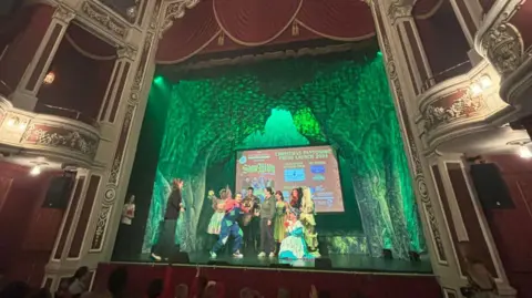 Stars gather on stage at the New Theatre Royal. The stage is lit green with trees as a backdrop, and you can see the royal boxes on the sides of the picture with classical style white and red paints