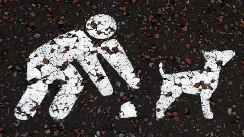 Getty Images Stencilled sign, painted in white, on a road depicts a dog owner leaning down to pick up dog faeces next to their pet.