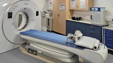 NHS Humber Health Partnership A large MRI scanner in a treatment centre room. A blue bed is in the centre of the room, attached to a large circular machine. There are blue cupboard doors around the edge of the room.