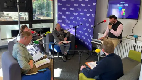 Four people sat on sofas in front of microphones. The presenter is sat on a chair. Behind him is purple BBC election branding.
