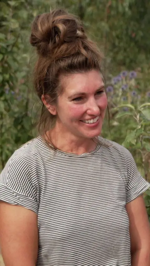A woman in a stripey top is smiling. She is standing outdoors in front of shrubs