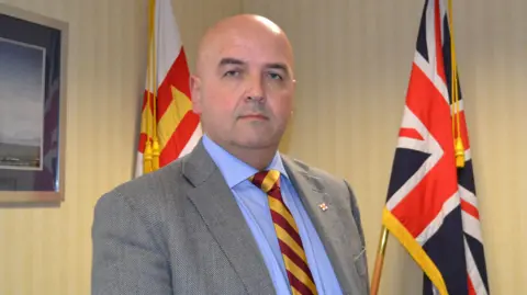 BBC A wide picture of Jonathan Le Tocq standing in front of two flags.