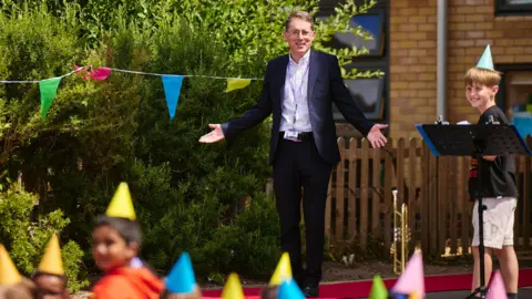 Wood Farm Primary School Mr Lewin stood with his arms out appearing very surprised, whilst a crowd of children watch on.