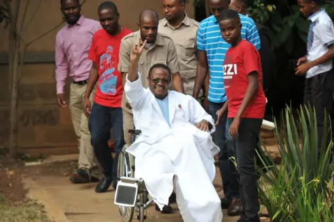 AFP Tundu Lissu waves from his wheelchair after a press conference surrounded by family and well-wishers at a hospital in Kenya's capital, Nairobi, on January 5, 2018.