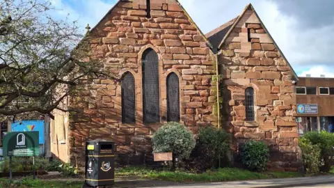 A red-brick building, which is home to Maggs Day Centre. Several bushes and a black bin are outside the building.