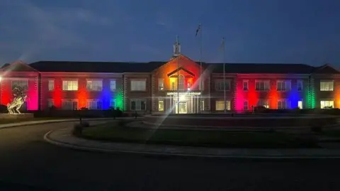 Dave Smith A two storey long brick building with a steeple in the evening lit by rainbow colours 
