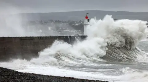 Greg Martin A huge foamy wave crashes into the pier at Newyln Harbour in Cornwall. The lighthouse can be seen beneath the wave, with a beach in the foreground and houses in the background