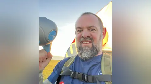 John Martin looking directly into the camera. He has short hair and a grey beard and is wearing a backpack while carrying a kettle bell in his right hand.