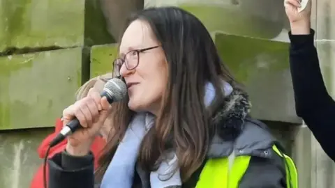 A woman with shoulder-length dark hair smiling as she speaks into a microphone at a rally. She is standing as part of a group in front of a sandstone building. A person wearing red can be glimpsed behind her and another person's raised arm can be seen to her right.