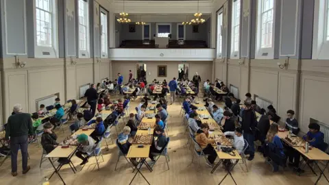 Shropshire Chess Association A hall full of wooden tables and chairs with people sitting at them playing chess. The floor is a pale wood and the walls are cream with large rectangular panelling. There is a balcony at the back of the room with dark, wooden seating, as well as large windows above the cream panels. 