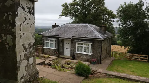 Crown Prosecution Service A small grey brick bungalow with white trim windows and a door, with a farmer's field and woods in the background. 