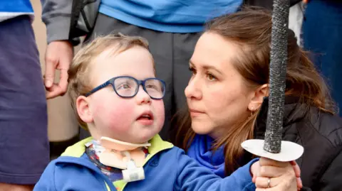 A child wearing glasses and holding a lantern. He is holding a toy sword and is stood next to his mother. 