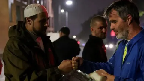 Reuters Adam Kelwick with a beard and green coat fist pumps a demonstrator with a grey beard, yellow t-shirt and blue coat and hands him food  