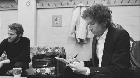 BDA Feinstein Black and white image of Dylan writing on a notepad in his hands as he sits in a room. Another man sits on a sofa to the left with a table and paper cups in between them.