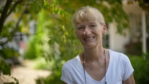 Jamie Niblock/BBC Fiona smiles at the camera, standing to the right of the shot. She is wearing a white T-shirt and has her hair tied up behind her head. She is standing in front of a tree is dappled sunlight. 