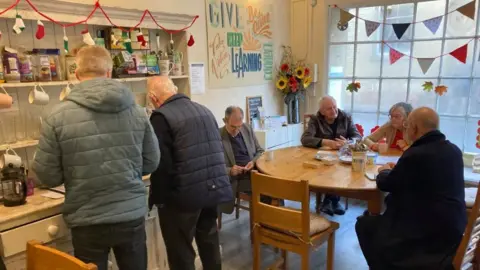 James Grant/BBC Six people sitting or standing around a table at a cafe