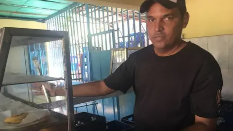 Luis Mendez stands at a café counter, wearing a black t-shirt and a baseball cap.