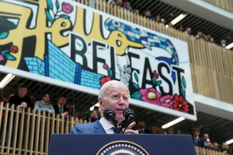 Reuters People watch as Joe Biden gives a speech beneath a large mural that reads: Hello Belfast