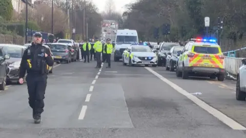 BBC Police officer approaches camera as numerous emergency vehicles line eaither side of street strung with crime scene tape