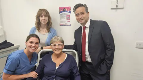 University Hospitals Birmingham A group of four people in a white hospital room, a woman in blue scrubs leans towards a woman in a blue top who is sitting in a hospital chair, behind them are a man in a grey suit with a red tie and a woman in a blue cardigan with a taupe top underneath