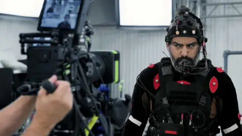 Sony A behind the scenes picture show Kohli taking part in a performance capture process, which is where an actor has their facial expressions and movements recorded for the game. He wears a black capture suit with red tabs, a helmet with a mouthpiece and has white dots on his face. A camera operator is seen to his left