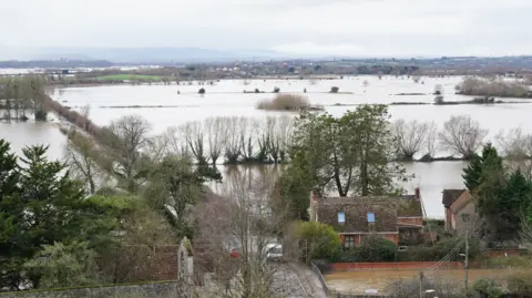 A number of flooded fields are seen from above. Two houses are visible in the foreground, surrounded by water. 