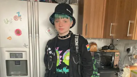Family handout Family photo of Jason Pulman in a kitchen. He wears a black t-shirt with a Rick and Morty illustration, a black jacket with several badges on it and a black bucket hat.