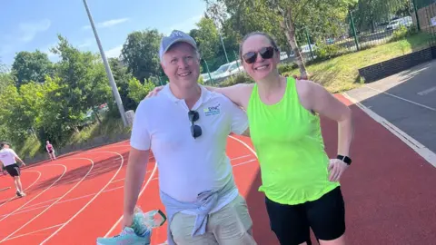 Sarah Dodd standing on the track of Wentworth Leisure Centre after her fundraising challenge. She is wearing her running clothes, including a bright green vest. She has her arms around a man, wearing a white shirt and cap.