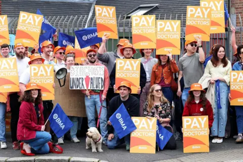 Pacemaker A large crowd of people on strike,, holding orange placards that read "pay restoration for doctors"