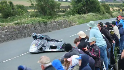 SOUTHERN 100 A sidecar outfit on the track with a stone wall on the left, and spectators leaning over another on on the right.
