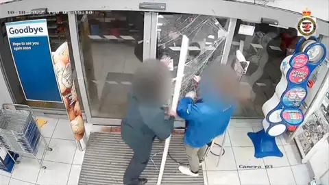 A CCTV video still shows two shopkeepers in a Tesco store standing in front of a glass door, at this time when an intruder is breaking the glass. 