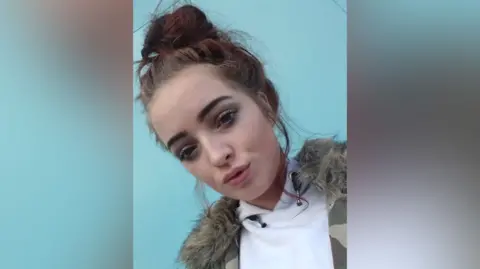 Family a young woman with dark brown hair worn in a top knot. She is wearing a white hooded top and a camouflage patterned coat with a fake fur collar