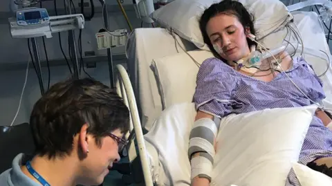 Becca Heritage Becca Heritage laying in a hospital bed, wearing a purple striped hospital down. She is attached to lots of wires and medical equipment, including a large tube attached to her throat. Her legs and arms have black patches on them. A female healthcare professional wearing an NHS lanyard is crouched by her bedside.